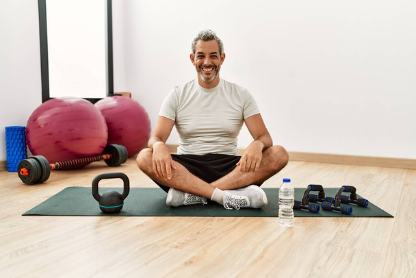 Hombre hispano de mediana edad sentado en una colchoneta de entrenamiento en el gimnasio luciendo positivo y feliz de pie y sonriendo con una sonrisa confiada mostrando los dientes  - Foto, Imagen