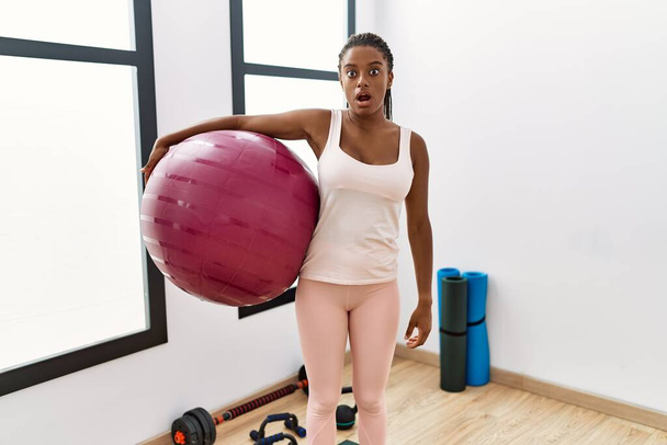 Jonge Afrikaans-Amerikaanse vrouw met vlechten houden pilate bal in yoga kamer bang en verbaasd met open mond voor verrassing, ongeloof gezicht  - Foto, afbeelding