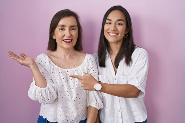 Hispanic mother and daughter together amazed and smiling to the camera while presenting with hand and pointing with finger.  - Photo, image