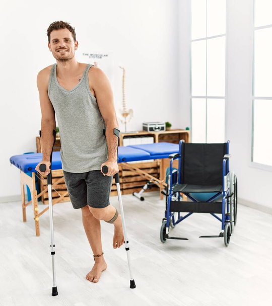 Young hispanic man smiling confident walking using crutches at clinic - Photo, Image