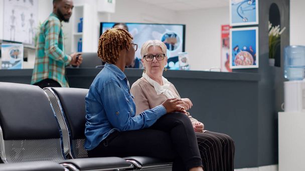 Senior patient and african american woman talking in waiting area at medical facility reception. Diverse people sitting in hospital waiting room to attend healthcare checkup visit. Handheld shot. - Fotó, kép