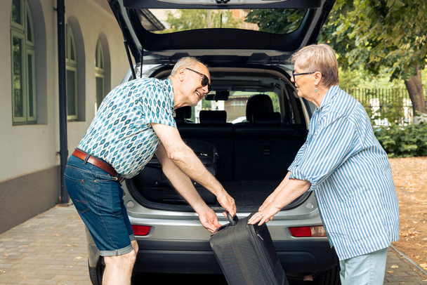 Senior people putting bags in vehicle trunk to leave on retirement holiday trip during summer. Travelling on road trip for recreation, packing baggage and suitcase for urban cityscape vacation. - Photo, Image