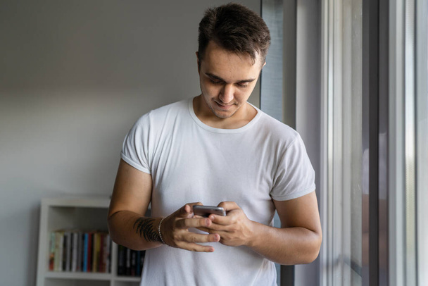 Ένας άνδρας νεαρός άνδρας καυκάσιος στέκεται δίπλα στο παράθυρο στο σπίτι φορούν λευκό πουκάμισο χρησιμοποιώντας το κινητό τηλέφωνο για να στείλετε sms μηνύματα ή περιήγηση στο διαδίκτυο μέσω της εφαρμογής κοινωνικό δίκτυο app εγκαταστήσετε χώρο αντίγραφο - Φωτογραφία, εικόνα