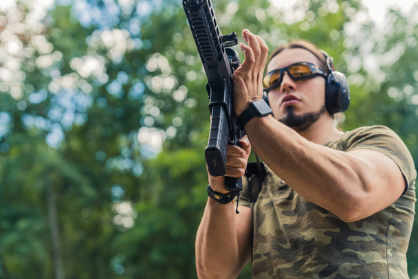 Caucasian bearded man in safety goggles and headphones wearing camo t-shirt reloading submachine gun. Outdoor horizontal shot. High quality photo - Photo, image