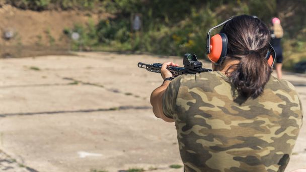 Rear view of man with ponytail wearing camo t-shirt and safety headphones practicing aim using submachine gun. Outdoor shooting range. Horizontal shot. High quality photo - 写真・画像
