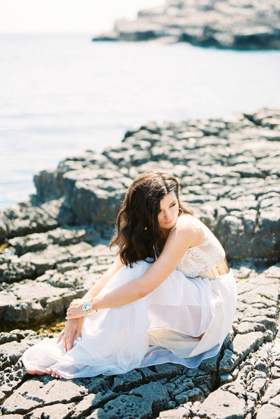 Босоногая невеста в белом платье сидит на камне, обнимая колени руками. Высокое качество фото - Фото, изображение