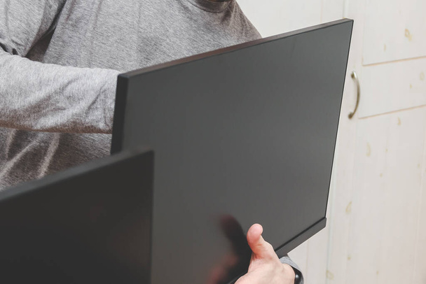 A young caucasian guy without a face holds a computer monitor with his hands, installing it on a bracket against the background of a blurry wardrobe in the room, side view close-up. The concept of repair at home. - 写真・画像