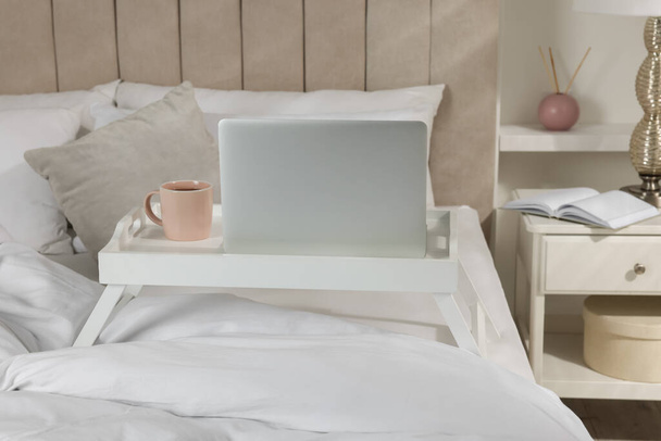 White tray table with laptop and cup of drink on bed indoors - Fotoğraf, Görsel