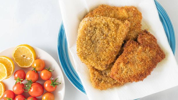 Crispy pan fried breaded pork chops served with lemon and fresh tomatoes close up on a plate on light grey background, flat lay - Valokuva, kuva