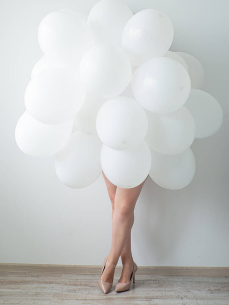 Женщина на бежевых высоких каблуках держит белые шарики на фоне белой стены. Ноги и много белых воздушных шаров. С днем рождения. Белое украшение. Копирование пространства - Фото, изображение