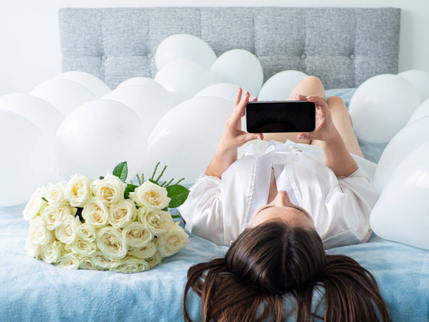 Женщина в белом лежит между белыми шариками на кровати. Портрет улыбающейся женщины с помощью телефона с большим количеством белых воздушных шаров и букета роз. С днем рождения, свадьба. Принято. Mock up. - Фото, изображение
