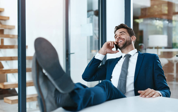 Making millions with one phone call. a handsome young businessman relaxing with his feet up on an office desk while using his phone - Zdjęcie, obraz
