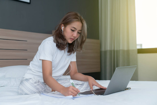 Beautiful Asian woman sitting and smiling the bed and her laptop. WFH Working from home in quarantine lockdown. Social distancing Self Isolation - Фото, зображення