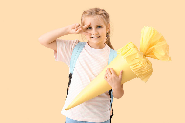 Cute little girl with yellow school cone showing victory gesture on beige background - Foto, Bild