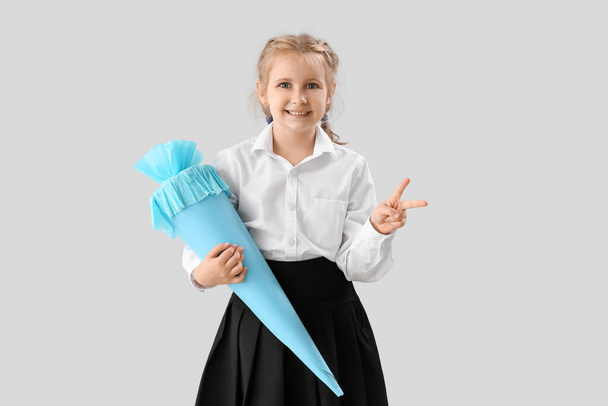 Cute little girl with blue school cone showing victory gesture on light background - Foto, Bild