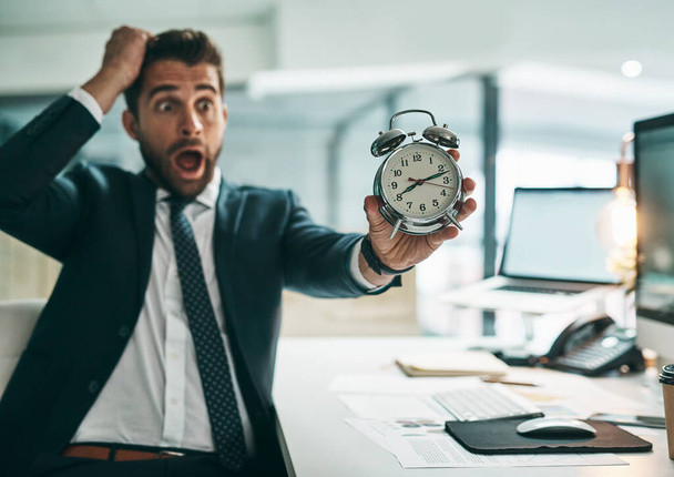 Oh dear is that the time already. a surprised young businessman holding an alarm clock while being seated in the office - Foto, imagen