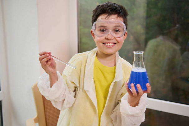 Happy, inspired schoolboy wearing lab coat and goggles smiles cutely at the camera while holding a glass flat-bottomed flask with combined blue chemicals during a science experiment in chemistry class - Fotó, kép