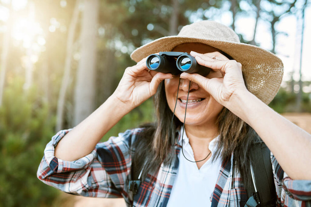 Female tourist hiking, looking through binoculars at wild birds in the trees. Happy, carefree and mature woman on nature walk, enjoying the view. Outdoor holiday time to promote health and wellness - Photo, image