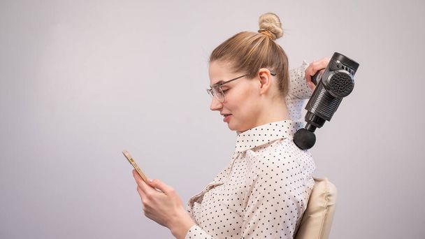 A young woman sits on a chair, uses a smartphone and makes herself massage with an electro massage gun - Fotoğraf, Görsel