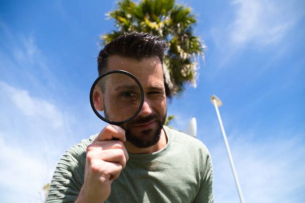 Handsome young man looks through a magnifying glass. His magnified eye can be seen from under the magnifying glass. The photo is taken from below and the man above. Concept detectives, view. - Foto, Bild
