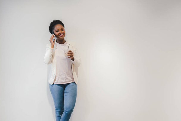 Young attractive african woman talking on her smart-phone in a coffee-shop, holding a glass of ice latte, leaning against white wall with copy space. Free space - Photo, Image