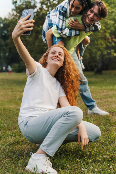 Red-haired girl taking a selfie with a boy who giving a piggyback to their girlfriend. Bloggers shooting stories for Instagram. - Фото, изображение