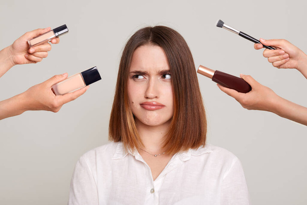 Indoor shot of confused puzzled woman preparing for party, deciding what make to do, looking away at cosmetics with pout lips, posing isolated on gray background. - Foto, imagen