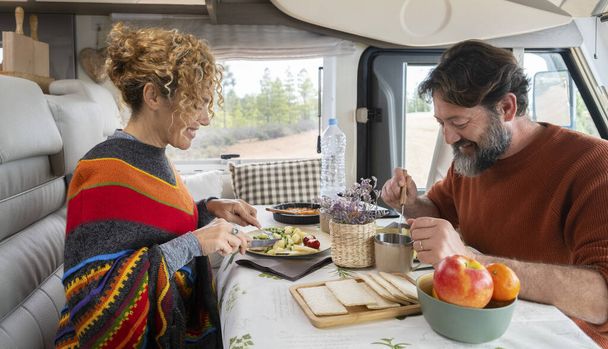 Young mature couple enjoy time together having lunch inside camper van. Happy people in free leisure holiday vacation with vehicle transport lifestyle. Alternative off grid house and freedom day - Photo, image