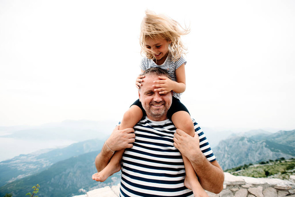 dad and daughter smiling and having fun together. daughter sitting on dad's shoulders and covers dad's eyes with hands. They are standing on the view point on Kotor bay, Montenegro. Wide angle photo - Photo, Image