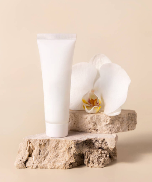 Weiße Cremetubus auf Travertinstein in der Nähe von weißen Orchideenblüten auf hellgelb, Nahaufnahme, Attrappe. Hautpflege handgemachtes Schönheitsprodukt, Creme oder Lotion. Exotische Naturkosmetik - Foto, Bild