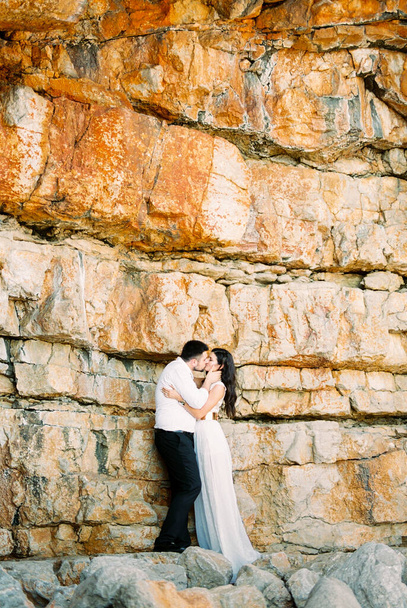 Groom kisses bride holding her face with his hands near a huge stone wall. High quality photo - Fotó, kép