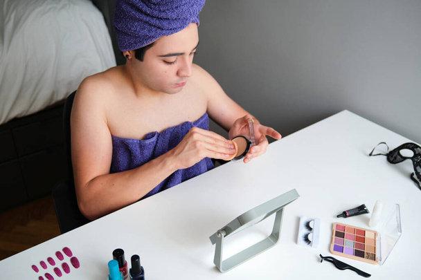 Young gender fluid person applying make-up base after shower with a towel on his head. - Foto, imagen