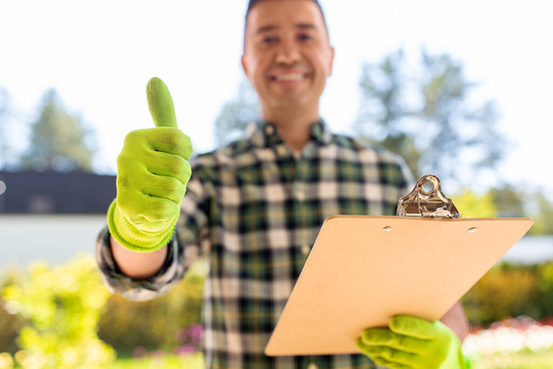 gardening and people concept - close up of happy smiling man with clipboard showing thumbs up at summer garden - Photo, Image