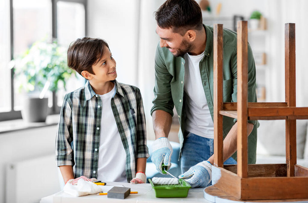 renovation, diy and home improvement concept - happy smiling father and son in gloves with paint roller painting old wooden table - Foto, afbeelding