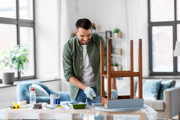 renovation, diy and home improvement concept - happy smiling man in gloves with paint roller painting old wooden table in grey color - 写真・画像