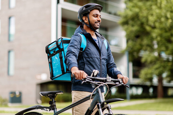 food shipping, profession and people concept - happy smiling indian delivery man in bike helmet with thermal insulated bag and bicycle on city street - Foto, immagini