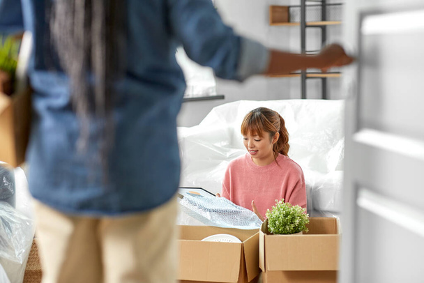 moving, people and real estate concept - woman unpacking boxes at new home and her friend entering room - Fotoğraf, Görsel