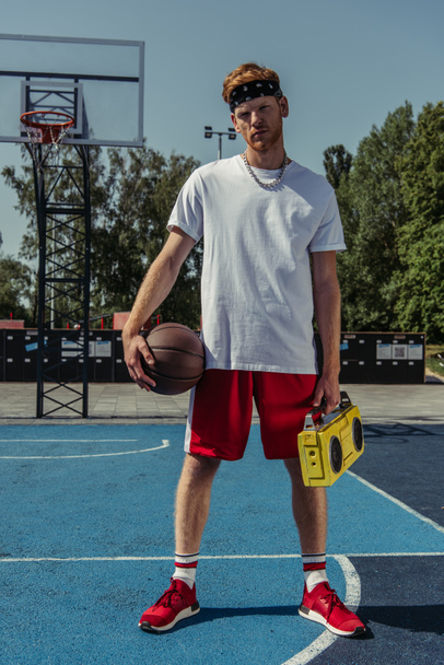 молодой баскетболист с мячом и бумбоксом смотрит в камеру на корте - Фото, изображение
