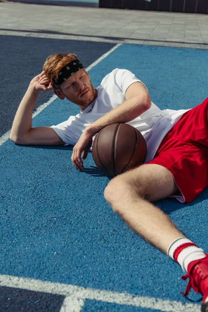 Рыжеволосый баскетболист в спортивной форме лежит на корте рядом с мячом и смотрит в сторону - Фото, изображение