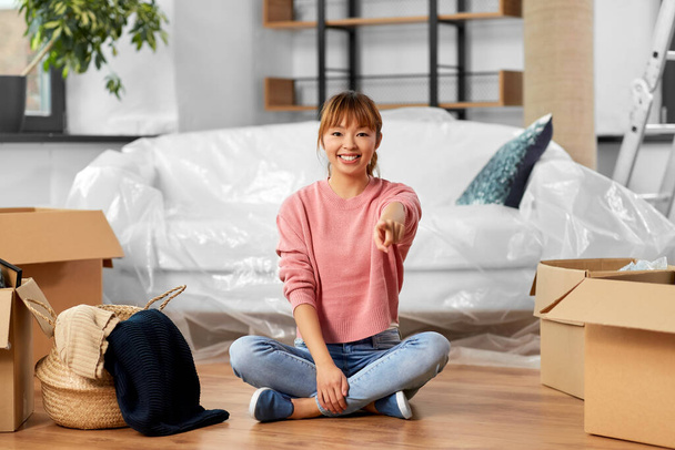 moving, people and real estate concept - happy smiling woman with boxes sitting on floor at new home and pointing to camera - Φωτογραφία, εικόνα