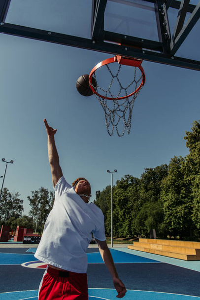 чоловік у білій футболці кидає м'яч у баскетбольне кільце на корті
 - Фото, зображення
