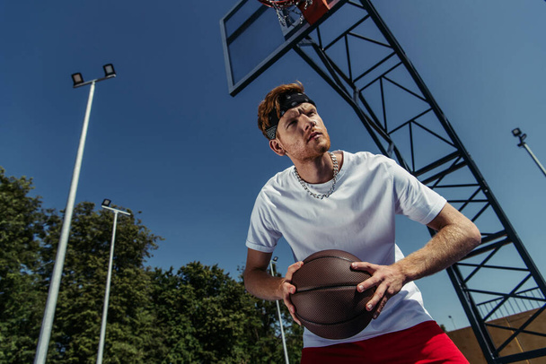 низкий угол обзора человека в бандане и белой футболке, играющей в баскетбол на открытом воздухе - Фото, изображение