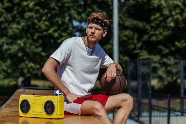 young basketball player sitting with ball near tape recorder and looking away - Photo, Image