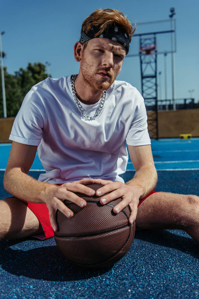 sportive man in headband sitting near ball on modern court - Photo, image