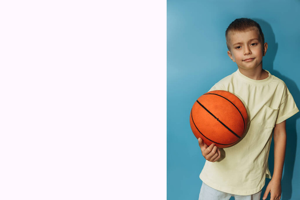 A serious boy of 6-7 years old in a yellow T-shirt on a blue background holds orange basketball. Portrait of a child, front view, white copy space - Foto, imagen