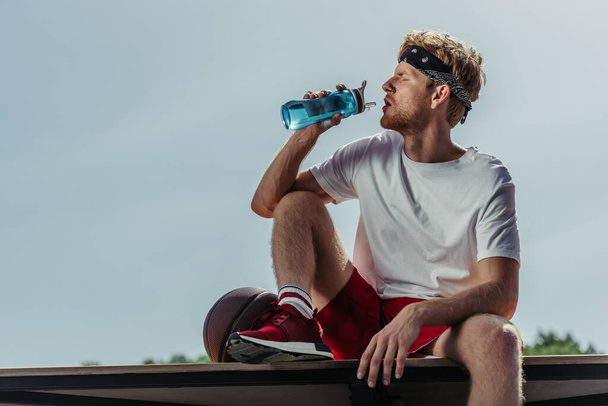 sportsman in white t-shirt drinking from sports bottle while sitting near ball - Photo, image
