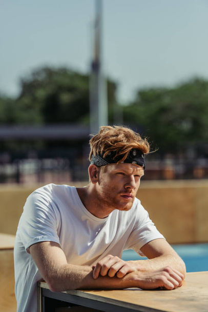 pensive redhead sportsman in white t-shirt and bandana looking away outdoors - Fotó, kép