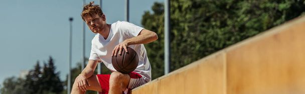 κοκκινομάλλα μπασκετμπολίστας κάθεται στο γήπεδο με μπάλα, λάβαρο - Φωτογραφία, εικόνα