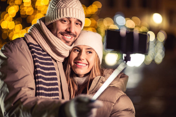 winter holidays and people concept - happy smiling couple taking picture with smartphone on selfie stick and hugging over christmas lights in evening - Foto, imagen