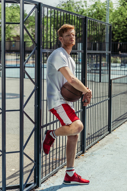 full length of man in sportswear standing with ball near fence of stadium - Zdjęcie, obraz